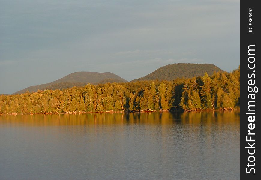 Golden sunlight at sunset on trees and twin mountains behind lake in Adirondacks, New York. Golden sunlight at sunset on trees and twin mountains behind lake in Adirondacks, New York.