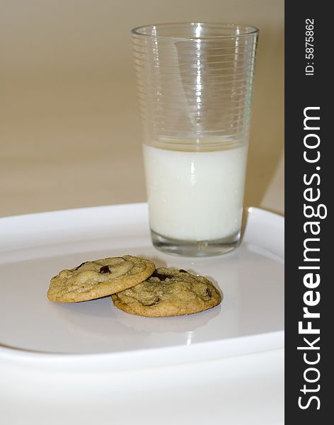 A pair of chocolate chip cookies sits stacked on a white surface, with a glass of milk in the background. A pair of chocolate chip cookies sits stacked on a white surface, with a glass of milk in the background.