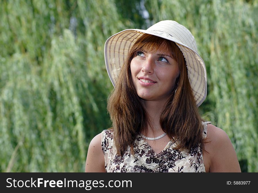 Beautiful smiling woman in the hat looks somewhere upward. A green tree branches on the smear background.