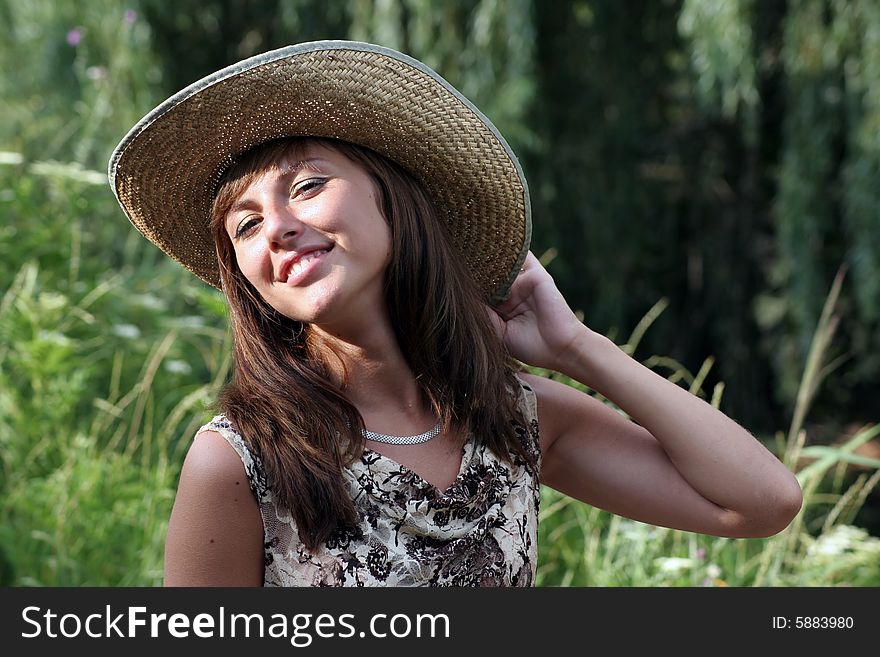 Coquettish woman in the strow hat smile at the camera. A green tree branches on the smear background.
