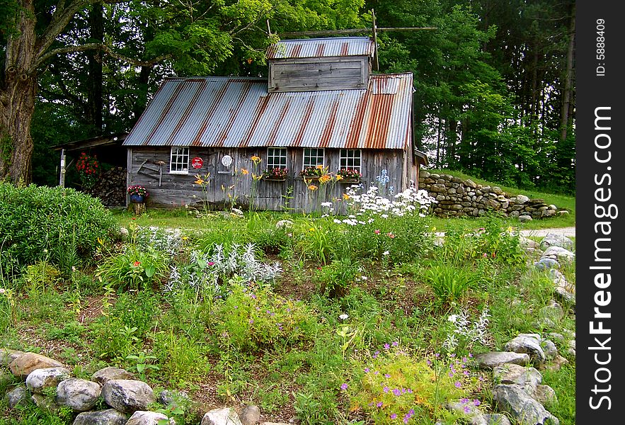 A beautiful sugar shack adorned with flowers in the window boxes for summer sits on a little hill surrounded with stone walls.