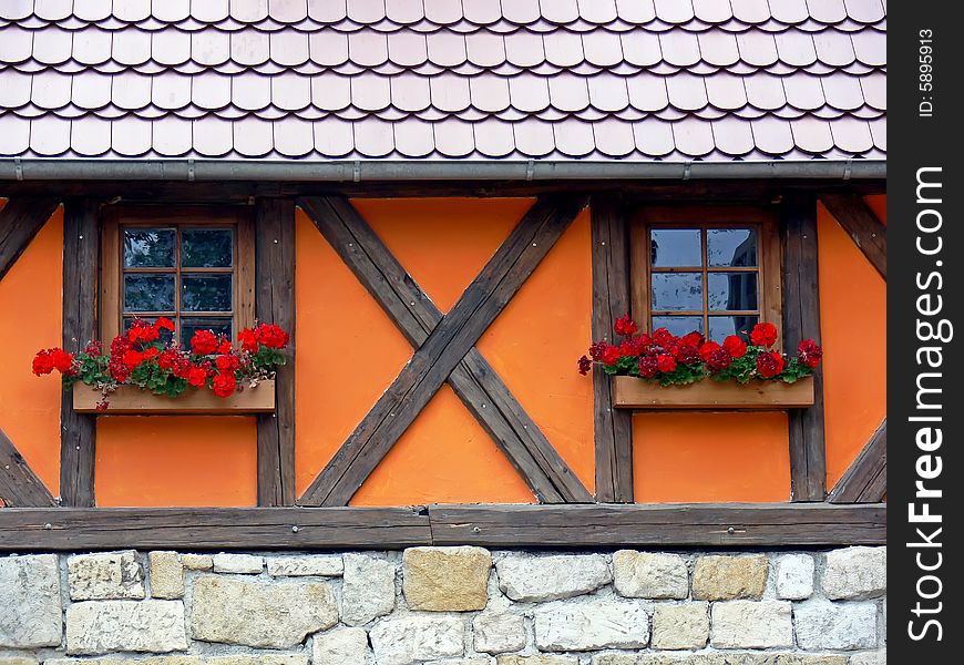 The old fashioned country home detail. Two wooden windows with red flowers in flower-pots. Post and pan, stone wall and new roof. The old fashioned country home detail. Two wooden windows with red flowers in flower-pots. Post and pan, stone wall and new roof.