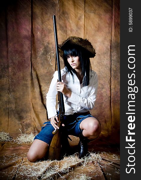 A young woman kneels holding an old shotgun. She wears a straw hat, white shirt and denim dungarees folded down. A young woman kneels holding an old shotgun. She wears a straw hat, white shirt and denim dungarees folded down.