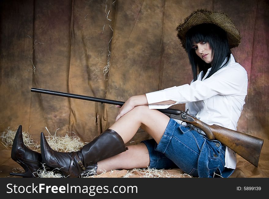 A young woman sits holding an old shotgun. She wears a straw hat, white shirt and denim dungarees folded down. A young woman sits holding an old shotgun. She wears a straw hat, white shirt and denim dungarees folded down.