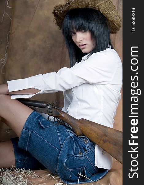 Portrait of a young lady with black hair and pink lipstick wearing denim skirt, white shirt and a straw hat. She is kneeling and holding an old shotgun. Straw is scattered around her feet. Portrait of a young lady with black hair and pink lipstick wearing denim skirt, white shirt and a straw hat. She is kneeling and holding an old shotgun. Straw is scattered around her feet.