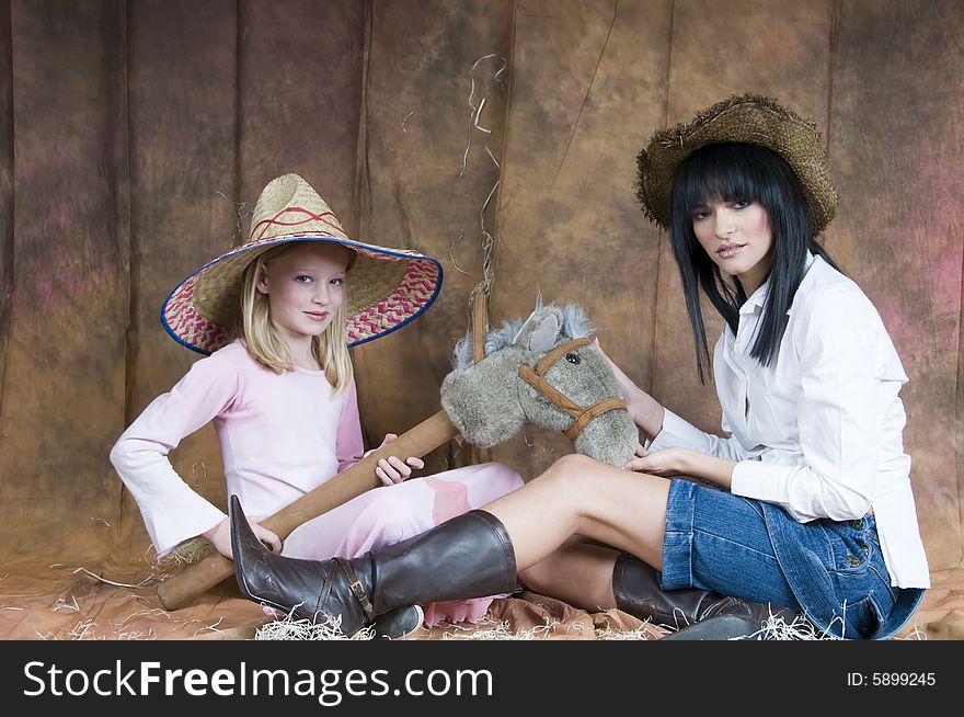 A landscape of a young woman in denim and white, and a young girl in a pink dress sit together wearing straw hats and playing with a toy horse. A landscape of a young woman in denim and white, and a young girl in a pink dress sit together wearing straw hats and playing with a toy horse.