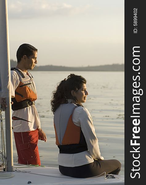 Man standing and woman sitting on bow of sailboat staring off into sunset. Vertically framed photo. Man standing and woman sitting on bow of sailboat staring off into sunset. Vertically framed photo.