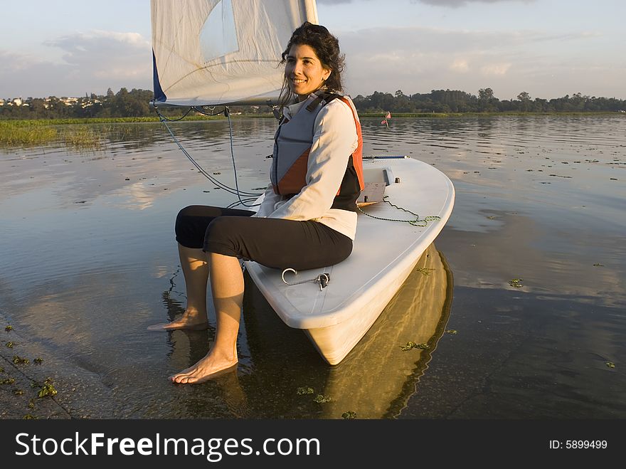 Woman sitting on bow of sailboat. Feet resting on ground in shallow water. Looking at camera and smiling. Horizontally framed photo. Woman sitting on bow of sailboat. Feet resting on ground in shallow water. Looking at camera and smiling. Horizontally framed photo.