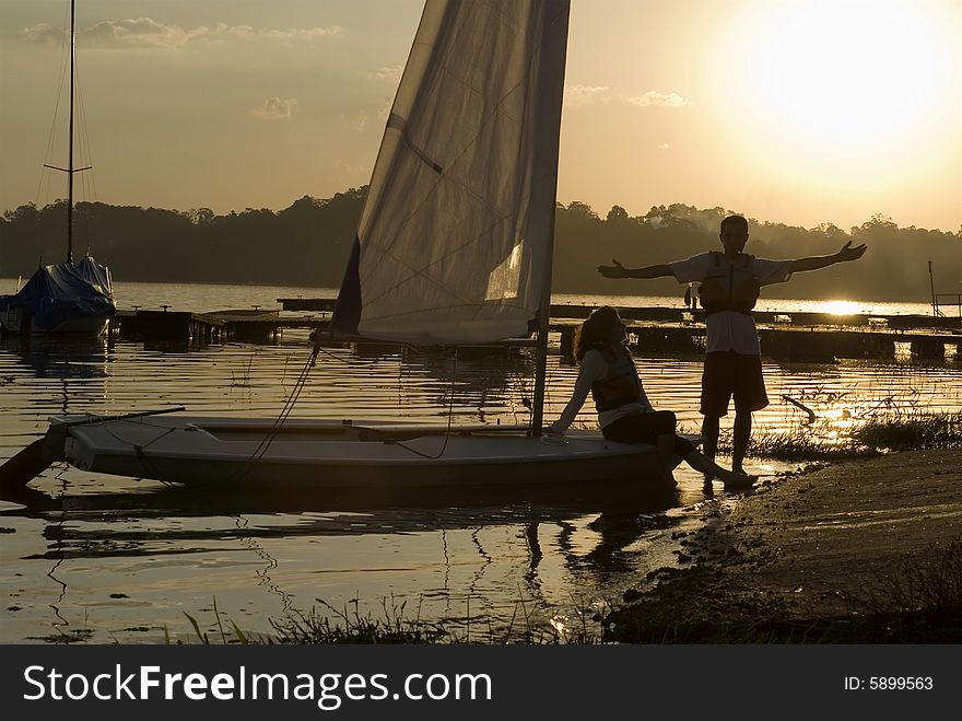 Woman sitting on sailboat. Man standing on shore in front of sunset. Horizontally framed photo. Woman sitting on sailboat. Man standing on shore in front of sunset. Horizontally framed photo