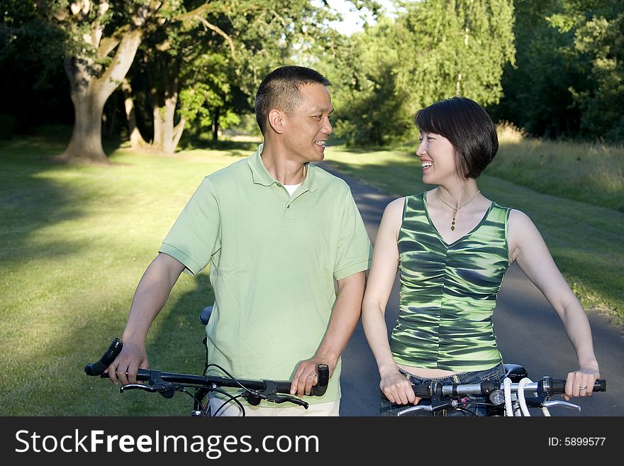 Asian couple standing alongside bicycles smiling at eachother. Couple standing in park. Horizontally framed photo. Asian couple standing alongside bicycles smiling at eachother. Couple standing in park. Horizontally framed photo.