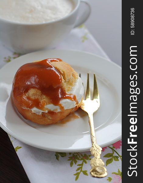 On a white plate one portion profiteroles with cream caramel sauce and a cup of cappuccino