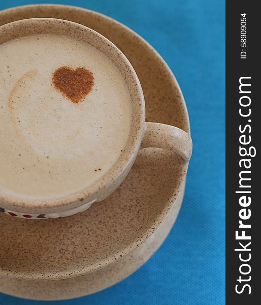 A cup of coffee with foam and cinnamon heart on a saucer and a bright blue napkin. A cup of coffee with foam and cinnamon heart on a saucer and a bright blue napkin