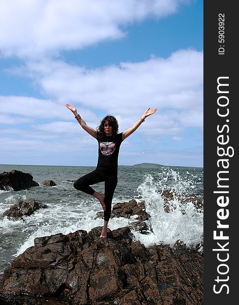 A beautiful woman doing yoga on the edge of the rocks with waves splashing to show a healthy way to live a happy and relaxed lifestyle in a world full of stress. A beautiful woman doing yoga on the edge of the rocks with waves splashing to show a healthy way to live a happy and relaxed lifestyle in a world full of stress