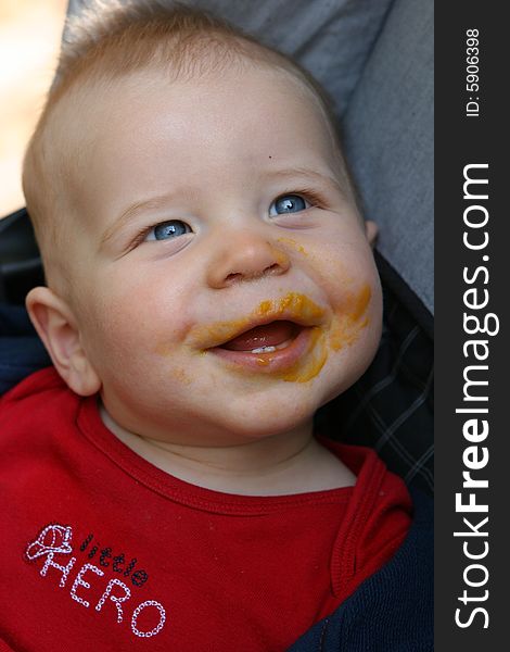 Newborn baby sitting and smiling with food on face.  The child has a red shirt and blue eyes. Newborn baby sitting and smiling with food on face.  The child has a red shirt and blue eyes