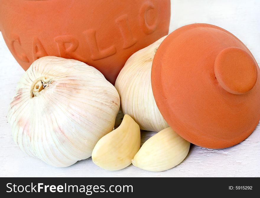 A clay garlic pot sits in front of fresh whole garlic and garlic cloves.  All on a worn white board. A clay garlic pot sits in front of fresh whole garlic and garlic cloves.  All on a worn white board.