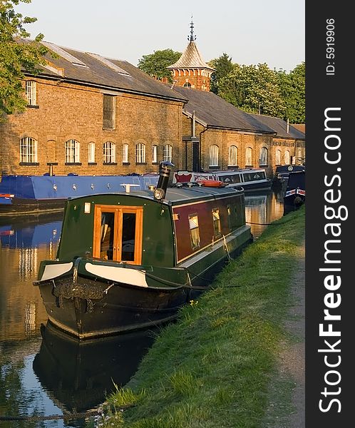 Narrow boats are reflected in the tranquil water of this British canal with a background of warm toned old buildings which are catching the evening sunshine. Narrow boats are reflected in the tranquil water of this British canal with a background of warm toned old buildings which are catching the evening sunshine.