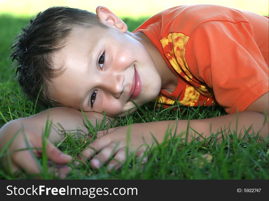 A little boy in a bright sport shirt lies on a green grass and smiles