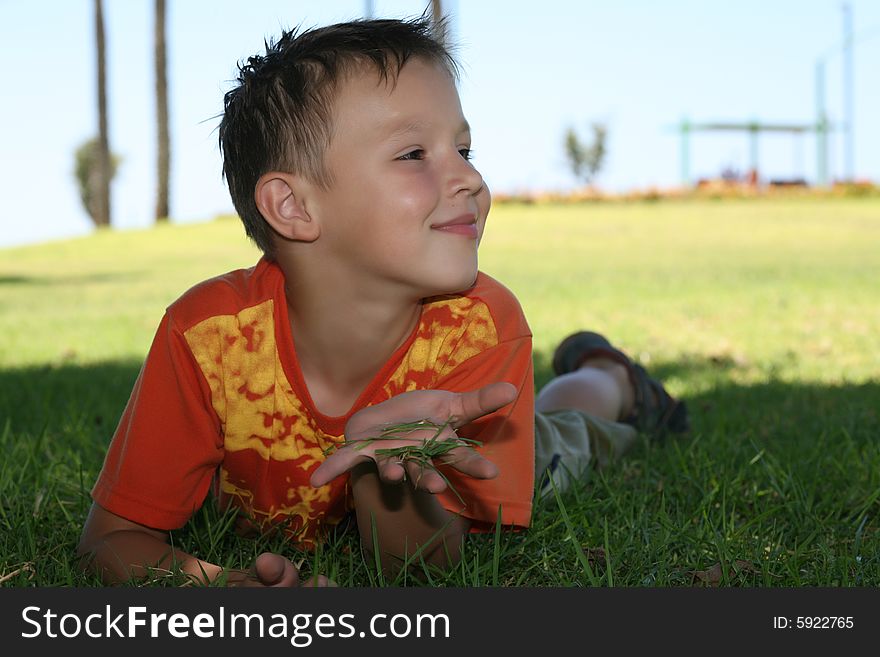 A little boy in an orange shirt lies and  holds a green  grass in a right arm