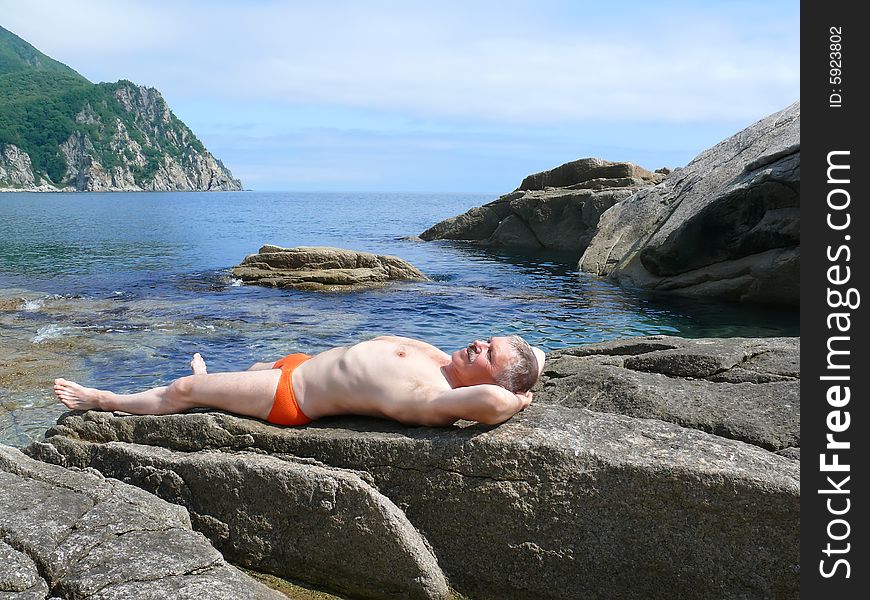 A man sunburns on stone at seacoast. On background are sea and blue sky with clouds and cape. Russian Far East, Primorye, Japanese sea. A man sunburns on stone at seacoast. On background are sea and blue sky with clouds and cape. Russian Far East, Primorye, Japanese sea.