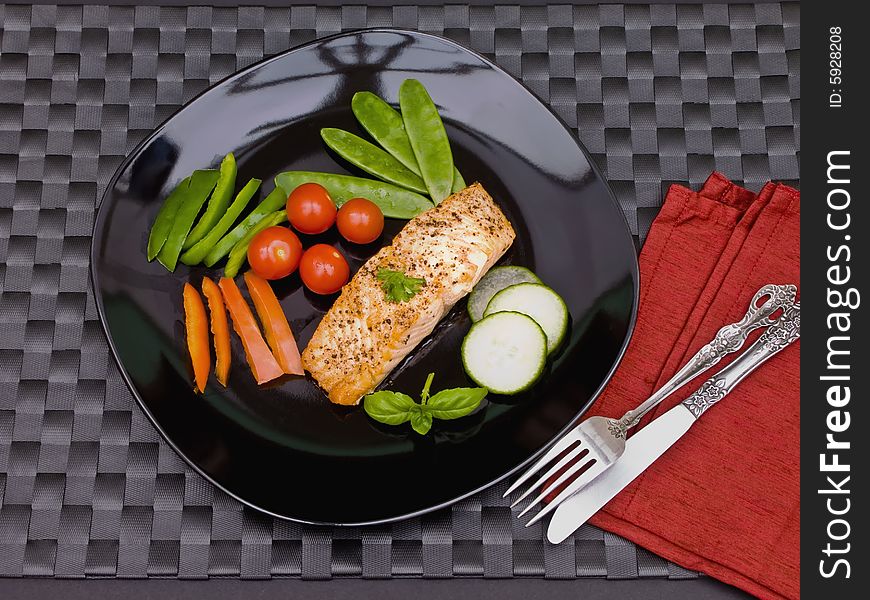 Salmon on black plate with cutlery and black mat.