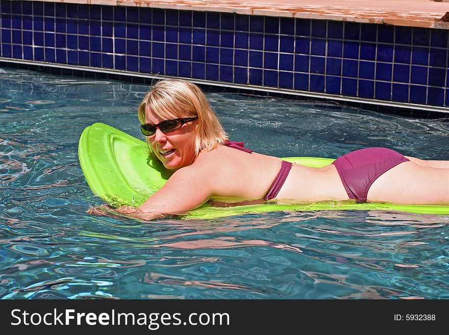 A beautiful blonde woman in a purple bikini floats on a raft in a swimming pool. She is wearing sunglasses as well. A beautiful blonde woman in a purple bikini floats on a raft in a swimming pool. She is wearing sunglasses as well.