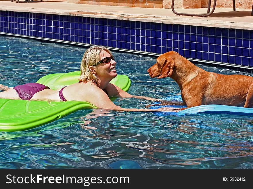 A beautiful blonde woman in a purple bikini floats on a raft in a swimming pool. She is facing a big red dog who is also floating on a raft. She is wearing sunglasses and has a big smile with white teeth. A beautiful blonde woman in a purple bikini floats on a raft in a swimming pool. She is facing a big red dog who is also floating on a raft. She is wearing sunglasses and has a big smile with white teeth.