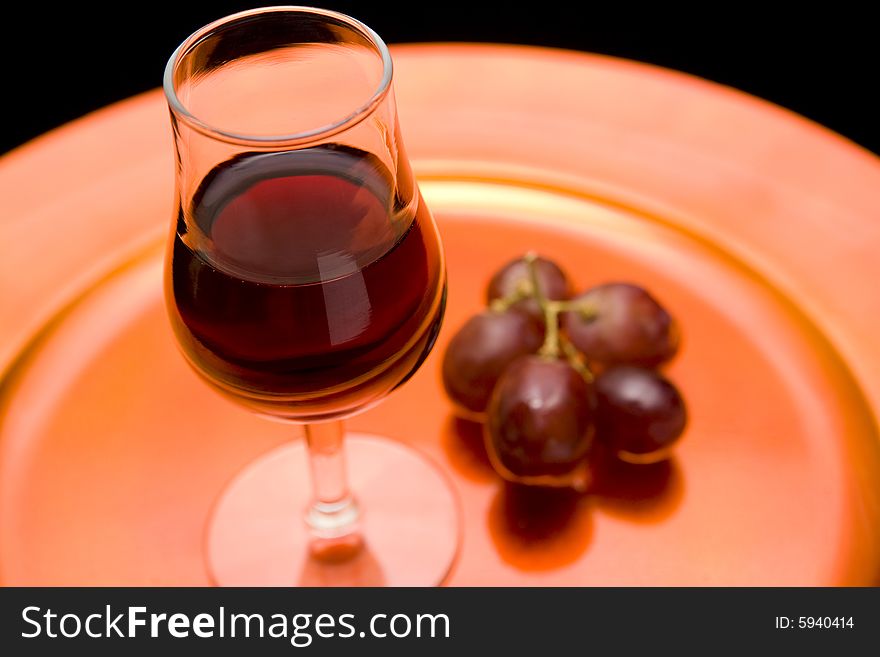 Closeup of a glass of red wine and red grapes on an orange tray. Black background. Closeup of a glass of red wine and red grapes on an orange tray. Black background