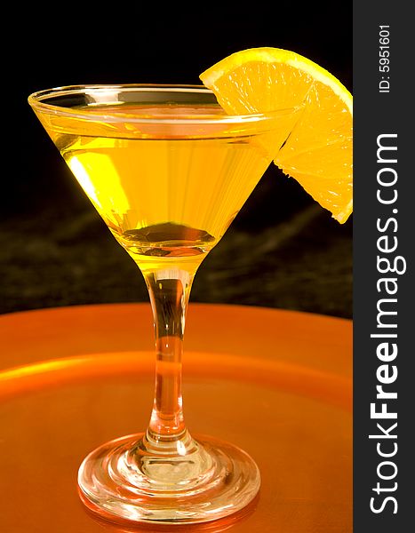 Closeup of a yellow cocktail on a bronze tray with a slice of an orange on a black background. Closeup of a yellow cocktail on a bronze tray with a slice of an orange on a black background