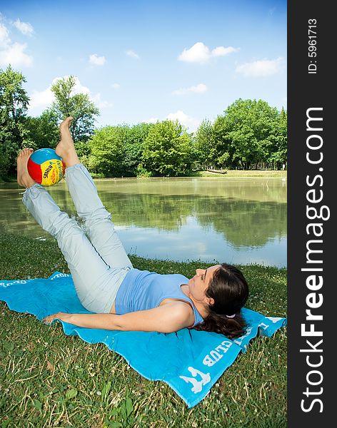 A girl, lying on a blue towel in the middle of the park near a little lake, doing some exercises with a ball. A girl, lying on a blue towel in the middle of the park near a little lake, doing some exercises with a ball