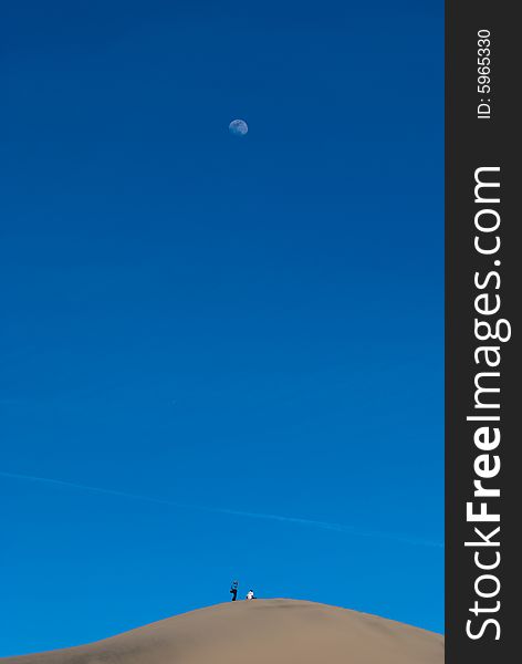 Two persons are standing on the top of a sand dunes with the moon above in the dark blue sky. Two persons are standing on the top of a sand dunes with the moon above in the dark blue sky.