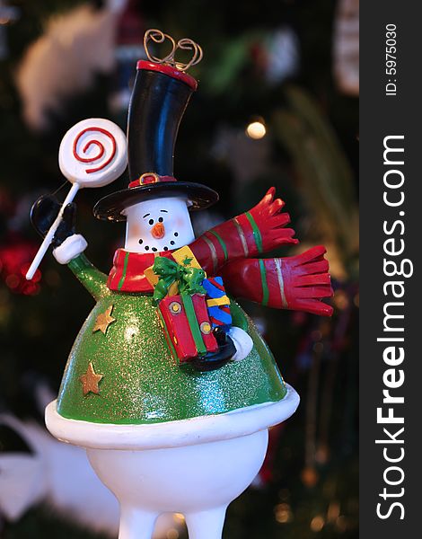 A decorative christmas snowman, holding gifts and waving, very blurry christmas tree in background. A decorative christmas snowman, holding gifts and waving, very blurry christmas tree in background