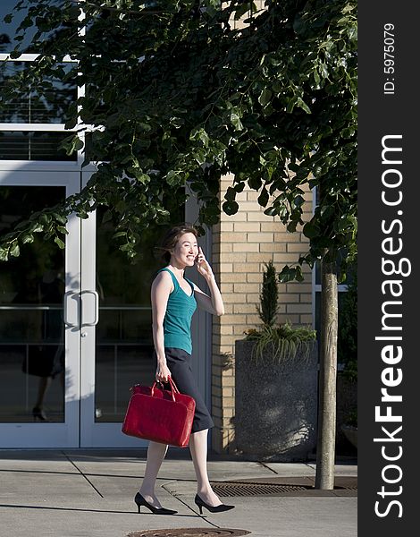 Young woman runs along building while talking on a cell phone. She is wearing a tank top and carrying a red bag. Vertically framed photo. Young woman runs along building while talking on a cell phone. She is wearing a tank top and carrying a red bag. Vertically framed photo.