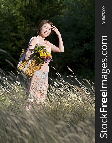 Attractive woman stands in a field while holding a basket containing a bouquet of flowers. She is standing to the side, smiling and brushing hair from her face.  Vertically framed photo. Attractive woman stands in a field while holding a basket containing a bouquet of flowers. She is standing to the side, smiling and brushing hair from her face.  Vertically framed photo.