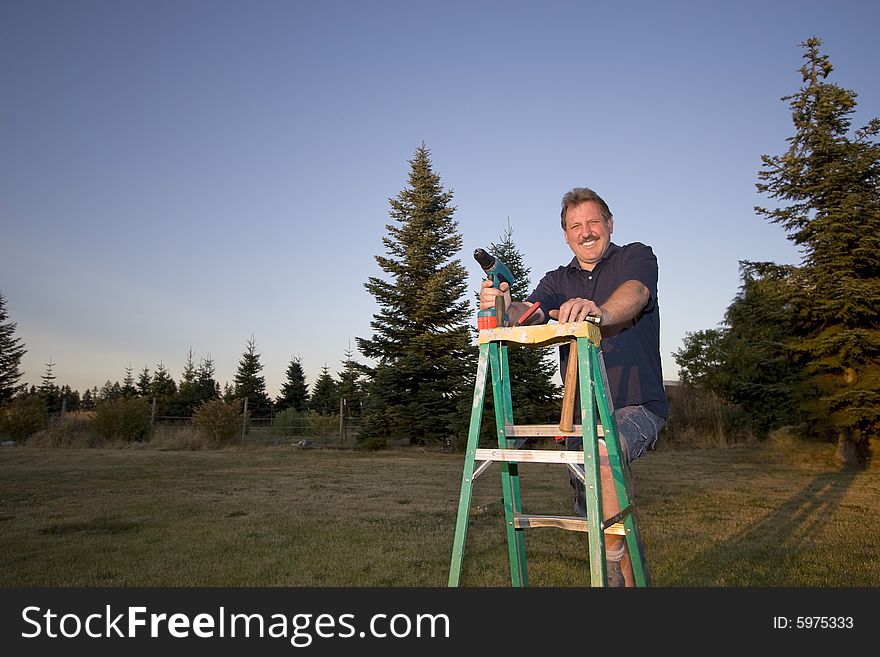 Man on ladder holding a drill and smiling. Horizontally framed photograph. Man on ladder holding a drill and smiling. Horizontally framed photograph