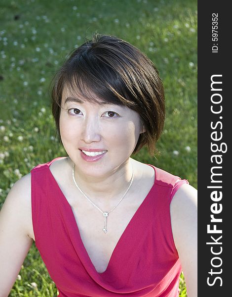 Young Asian woman smiles at camera while sitting on grass. Vertically framed photo. Young Asian woman smiles at camera while sitting on grass. Vertically framed photo.