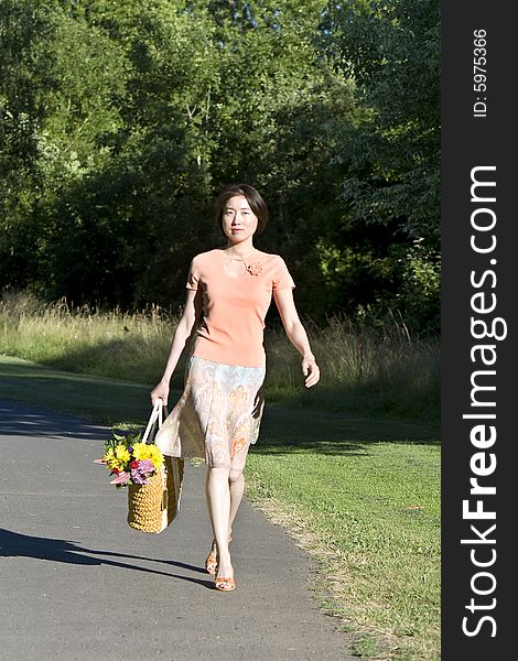 Attractive woman walks down path in a park carrying a basket containing a bouquet of flowers. Vertically framed photo. Attractive woman walks down path in a park carrying a basket containing a bouquet of flowers. Vertically framed photo.