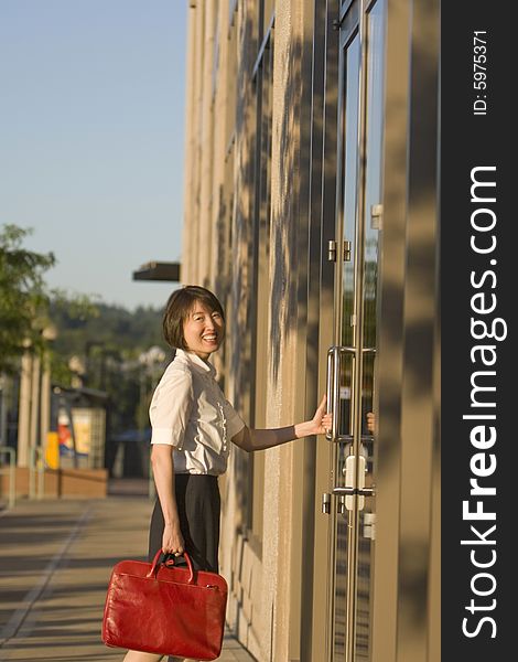 Young woman smiles at camera while opening a door to a building. She is smiling at the camera and carrying a red bag. Vertically framed photo. Young woman smiles at camera while opening a door to a building. She is smiling at the camera and carrying a red bag. Vertically framed photo.