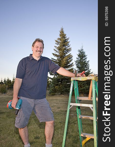 Man standing next to a ladder holding a drill and smiling. Vertically framed photograph. Man standing next to a ladder holding a drill and smiling. Vertically framed photograph