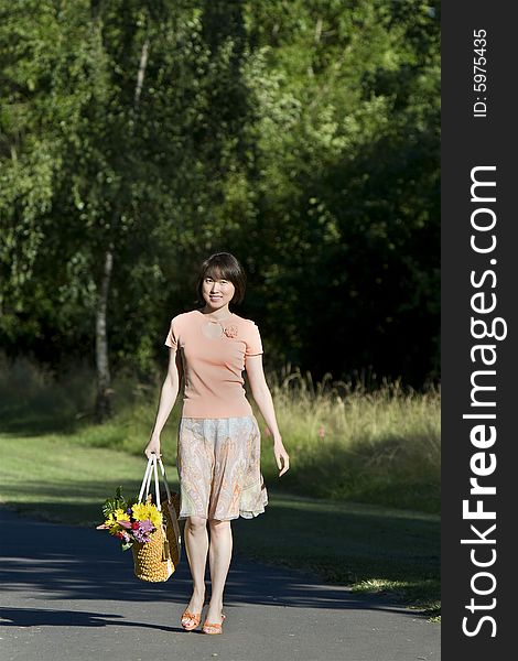 Attractive woman walks down path while carrying a basket containing a bouquet of flowers. She is smiling at camera.  Vertically framed photo. Attractive woman walks down path while carrying a basket containing a bouquet of flowers. She is smiling at camera.  Vertically framed photo.