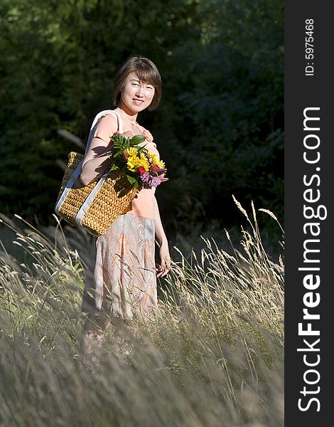 Attractive woman stands in a field while holding a basket containing a bouquet of flowers. She is standing to the side, smiling.  Vertically framed photo. Attractive woman stands in a field while holding a basket containing a bouquet of flowers. She is standing to the side, smiling.  Vertically framed photo.