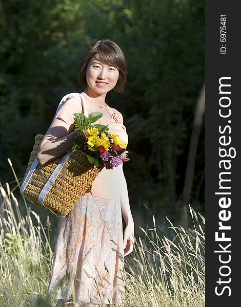 Attractive woman stands in a field while holding a basket containing a bouquet of flowers. She is standing to the side and smiling at the camera.  Vertically framed photo. Attractive woman stands in a field while holding a basket containing a bouquet of flowers. She is standing to the side and smiling at the camera.  Vertically framed photo.