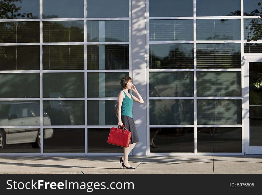 Young woman walks along building while talking on a cell phone and carrying a red bag. She is wearing a tank top. Horizontally framed photo. Young woman walks along building while talking on a cell phone and carrying a red bag. She is wearing a tank top. Horizontally framed photo.