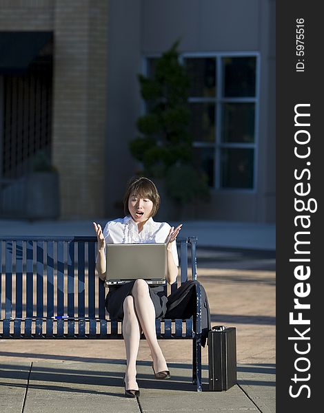 Young woman sits on a bench in front of a building. She is holding her laptop and looks surprised. Vertically framed photo. Young woman sits on a bench in front of a building. She is holding her laptop and looks surprised. Vertically framed photo.