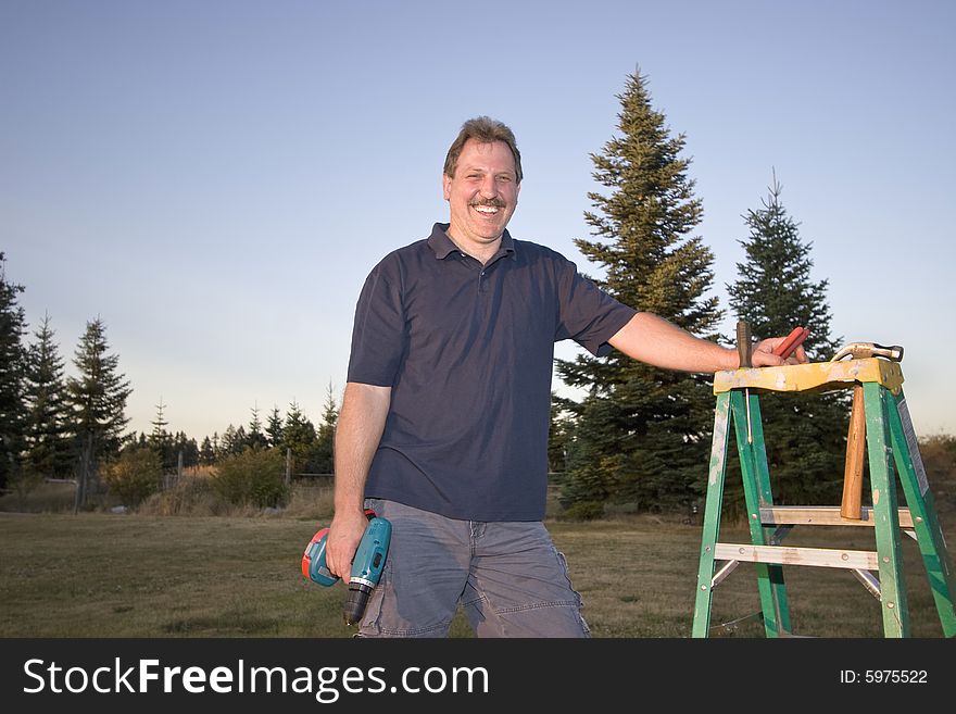 Man standing next to a ladder holding a drill and smiling. Horizontally framed photograph. Man standing next to a ladder holding a drill and smiling. Horizontally framed photograph