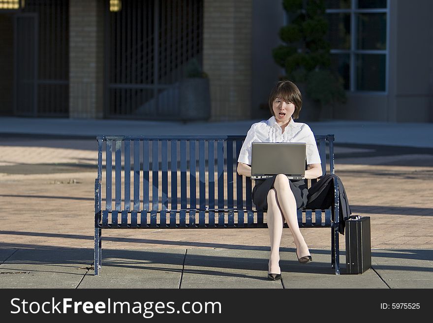 Young woman sits on a bench in front of a building. She is looking at her laptop and looks surprised. Vertically framed photo. Young woman sits on a bench in front of a building. She is looking at her laptop and looks surprised. Vertically framed photo.