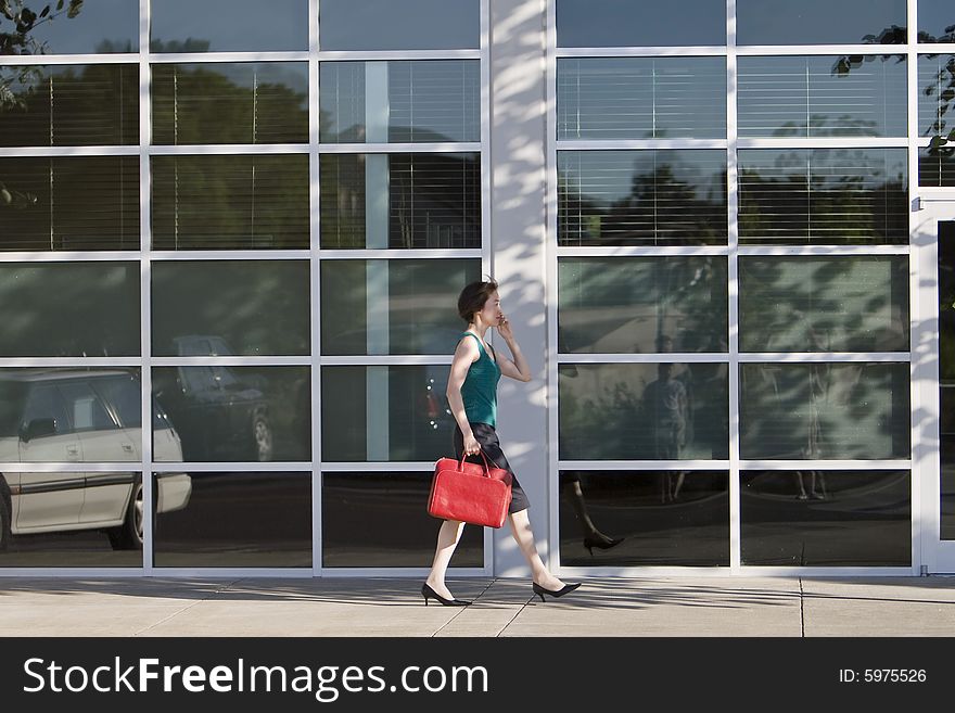 Young woman walks along building while talking on a cell phone and carrying a red bag. She is wearing a tank top. Horizontally framed photo. Young woman walks along building while talking on a cell phone and carrying a red bag. She is wearing a tank top. Horizontally framed photo.