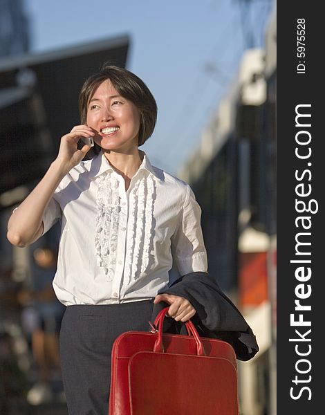 Young woman walks while talking on her cell phone. She is carrying a red bag. Vertically framed photo. Young woman walks while talking on her cell phone. She is carrying a red bag. Vertically framed photo.