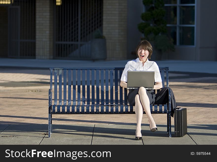 Young woman sits on a bench in front of a building. She is holding her laptop, looking surprised at the camera. Vertically framed photo. Young woman sits on a bench in front of a building. She is holding her laptop, looking surprised at the camera. Vertically framed photo.