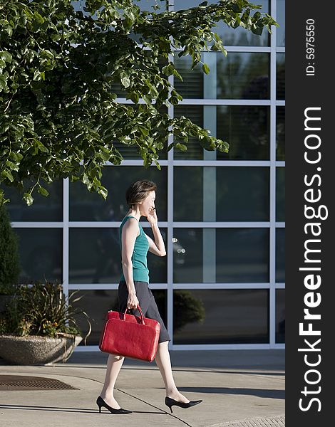 Young woman runs along building while talking on a cell phone. She is wearing a tank top and carrying a red bag. Vertically framed photo. Young woman runs along building while talking on a cell phone. She is wearing a tank top and carrying a red bag. Vertically framed photo.