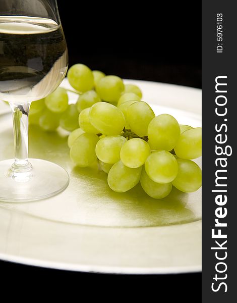 White wine and white grapes on a silver tray. Black background. Focus on grapes. White wine and white grapes on a silver tray. Black background. Focus on grapes